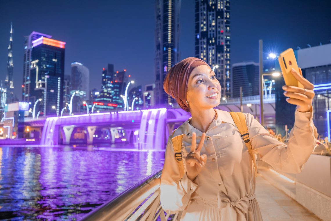Happy woman blogger takes a beautiful selfie photo against the background of an artificial waterfall illuminated in neon light from a bridge in downtown Dubai. Social networks and travel in the UAE