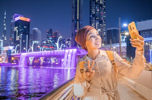 Happy woman blogger takes a beautiful selfie photo against the background of an artificial waterfall illuminated in neon light from a bridge in downtown Dubai. Social networks and travel in the UAE