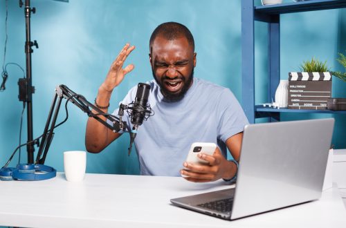Furious african american blogger reading negative feedback on smartphone while recording video. Angry upset vlogger holding mobile phone and sharing bad news while streaming live